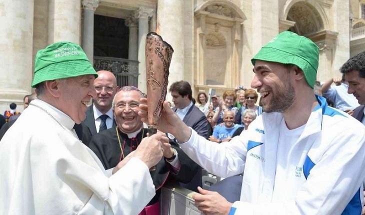 Na imagem, o papa francisco está com um chapéu verde igual a de um homem na multidão que mostra ao papa sorridente um objeto característico de algum lugar. Dia do papa.