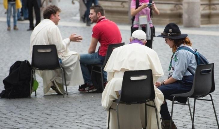 Na imagem, o papa francisco está sentado em uma cadeira simples na praça do vaticano com outros membros da igraja realizando confissões no povo. Dia do Papa.