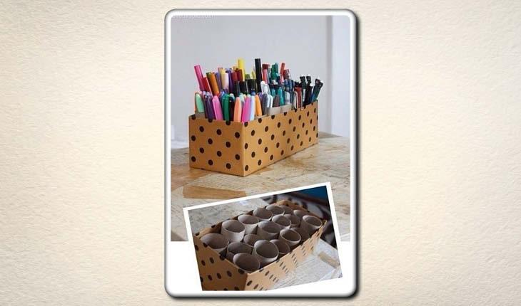 Na imagem há uma caixa de sapato encapada com papel laranja de bolinhas pretas sendo utilziada para organizar lápis e canetas. Dentro da caixa há rolos de papel higiênico em que as canetas são colocadas.