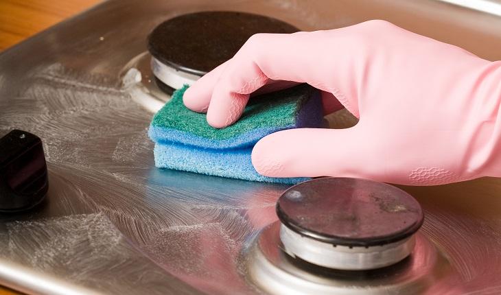 Mão de mulher limpando fogão com luva colorida na cor rosa e uma esponja de cozinha azul.