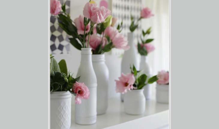 Na foto há diversas garrafas de tamanhos diversos pintadas de branco e dentro de cada uma delas há flores rosa-claro, como rosas.