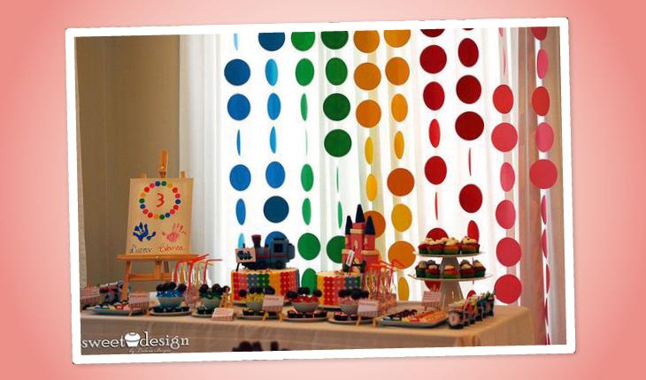 Na imagem há uma ideia de decoração de festa infantil e o fundo da imagem é rosa-claro. Na foto é possível ver a mesa do bolo e seu fundo com uma decoração feita de círculos de papel colorido pendurados.