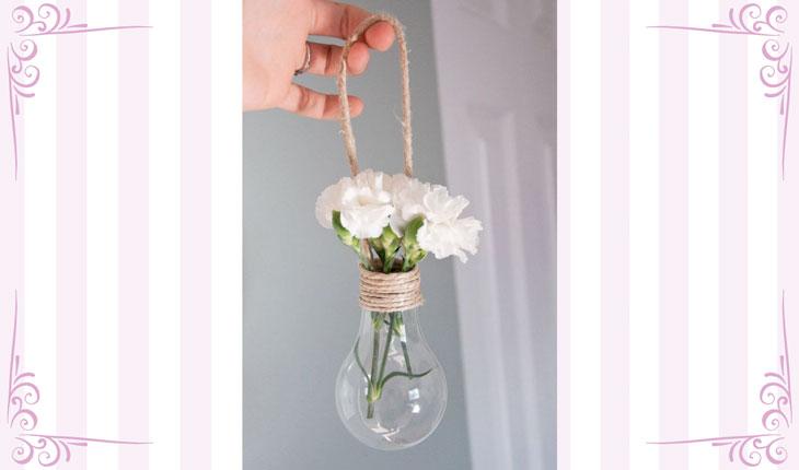 Na imagem há uma foto centralizada de um enfeite feito com uma lâmpada velha com flores dentro da abertura. Para pendurar, é utilizada uma corda. Ao redor há uma moldura branca com detalhes em rosa.