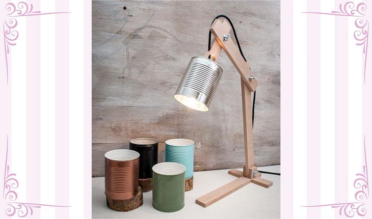 Na imagem há uma foto centralizada de uma luminária feita com latas de aço pintadas. O suporte é de madeira e a lâmpada fica dentro da lata. A luz está acesa. Ao redor há uma moldura branca com detalhes em rosa.
