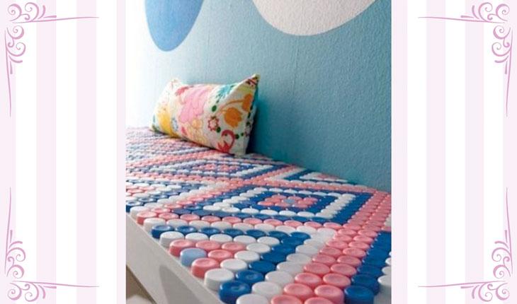Na imagem há uma foto centralizada de um banco branco revestido com tampinhas de garrafa nas cores azul, branca e rosa. Em cima do banco há uma almofada.