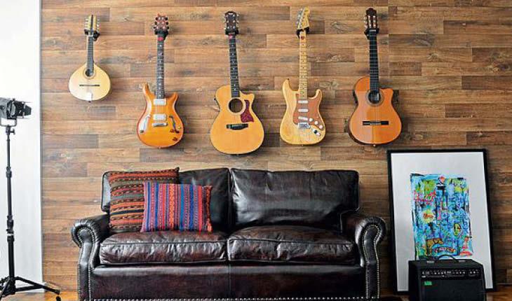 Na foto há uma sala com um sofá escuro e um painel de madeira atrás. Pendurados em cima do sofá estão violões e guitarras como decoração.
