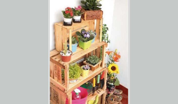 Na foto há diversos caixotes de madeira empilhados formando uma pequena estante. Dentro delas, há vasinhos coloridos com plantas.