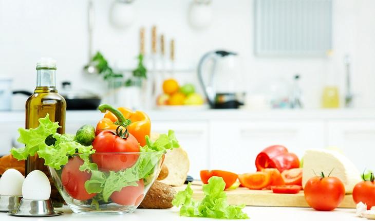 Tomates, ovos, queijo, alface, pimentão, abobrinha, e azeite em cima de uma bancada de cozinha branca. Os legumes estão entro de um bowl de vidro.
