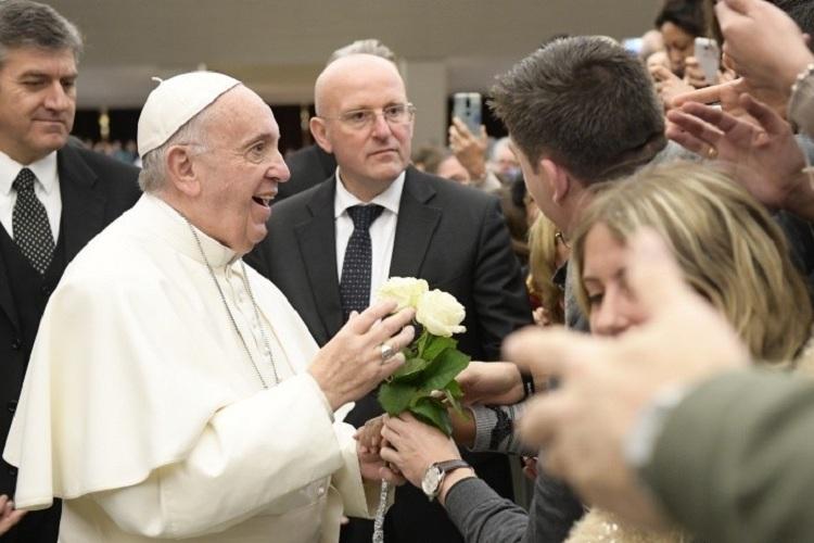 Na imagem, o papa francisco está recebendo flores dos fiéis e sorrindo bastante com uma multidão olhando para ele e tentando falar com ele. Mensagem de amor.