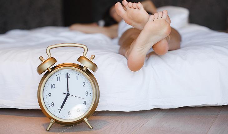 A imagem mostra uma mulher deitada em uma cama com um despertador perto dos seus pés. Fazer pequenas pausas para descanso ajuda você a ficar mais focada e produtiva