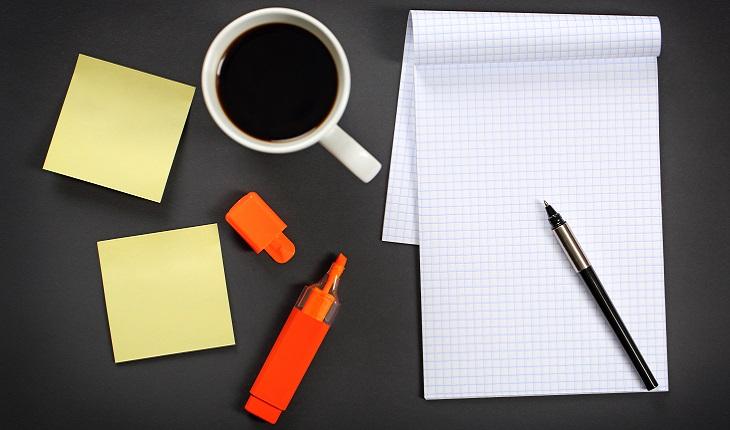 A foto mostra uma mesa com uma xícara de café, papel, lápis e marcadores. Organizar tarefas diárias é uma ótima maneira de se manter focada e produtiva