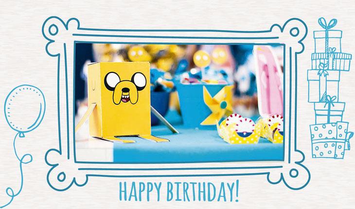 Na imagem há a foto da mesa do bolo, focando no papertoy do Jake, um dos personagens principais. Ao redor há uma moldura branca com desenhos em azul temáticos de festa de aniversário.