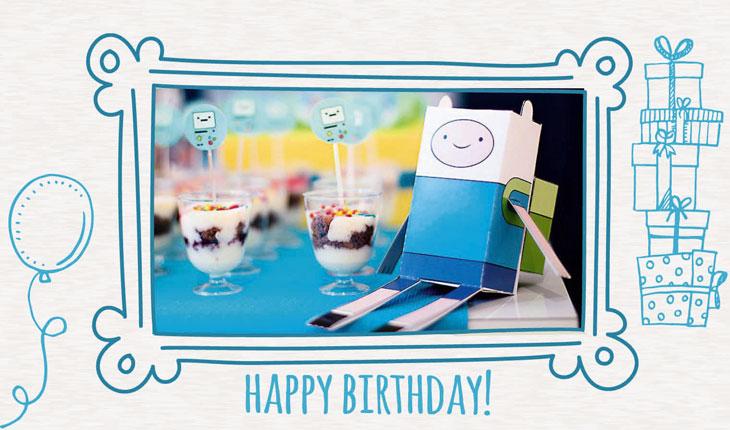 Na imagem há uma foto da mesa do bolo focando no papertoy do Finn, personagem principal do desenhos Hora da Aventura. Ao redor há uma moldura branca com desenhos azuis com a temática de festa de aniversário