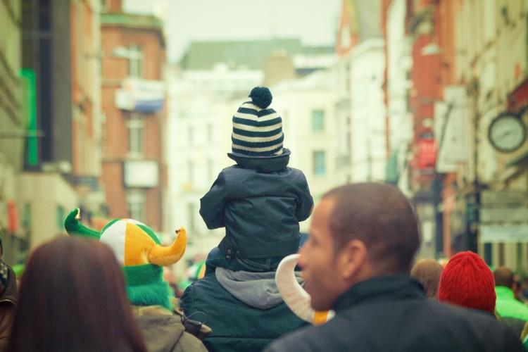 Imagem uma pessoa carregando uma criança nos ombros em meio a multidão, de costas para a câmera. Proteção para o amor e a família