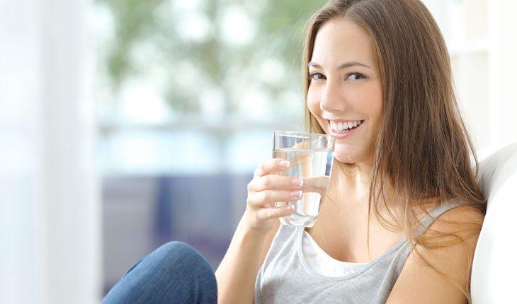 Unhas Impecáveis. Na foto, uma mulher segurando um copo d'água e sorrindo. Ela está sentada, com os cabelos solto, usando uma blusa cinza e uma calça jeans