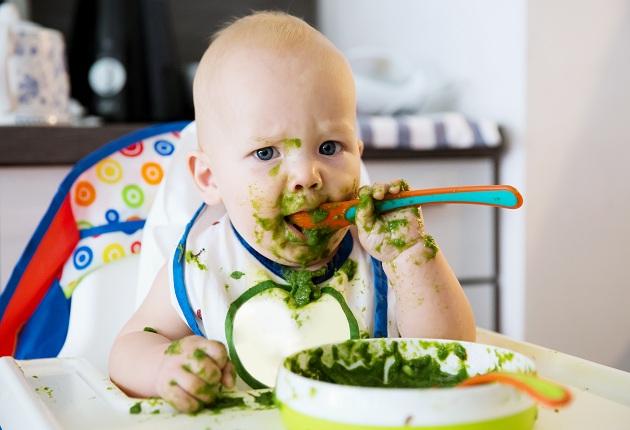 Bebê sujo sentado numa cadeirinha comendo papinha verde