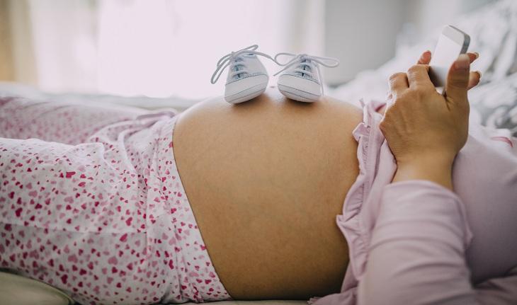 Imagem de uma mulher grávida deitada, vestindo pijama e com um par de tênis de bebê sobre a barriga. Gravidez