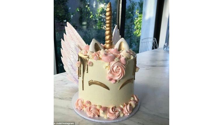 Na foto há um bolo redondo de unicórnio em que o bolo forma o rosto do unicórnio. Na base do bolo há detalhes de flores rosas. Os detalhes do rosto são dourados, assim como o chifre. A crina é rosa-claro também. Na parte detrás há duas asas rosa-claro. O rosto é feito apenas do os olhos fechados do unicórnio.