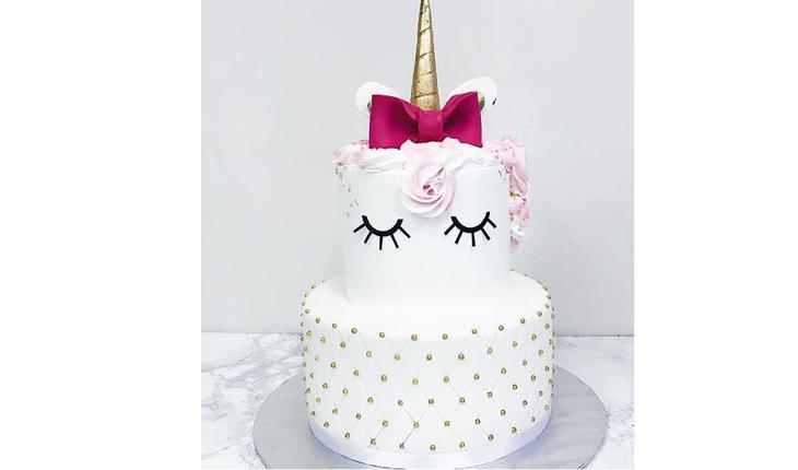 Na foto há um bolo de dois andares branco. O bolo de base tem bolinhas rosas em toda a volta. O bolo de base tem os olhos do unicórnio desenhados em preto. A crina é rosa, o chifre é dourado e há um laço pink ao redor do chifre.