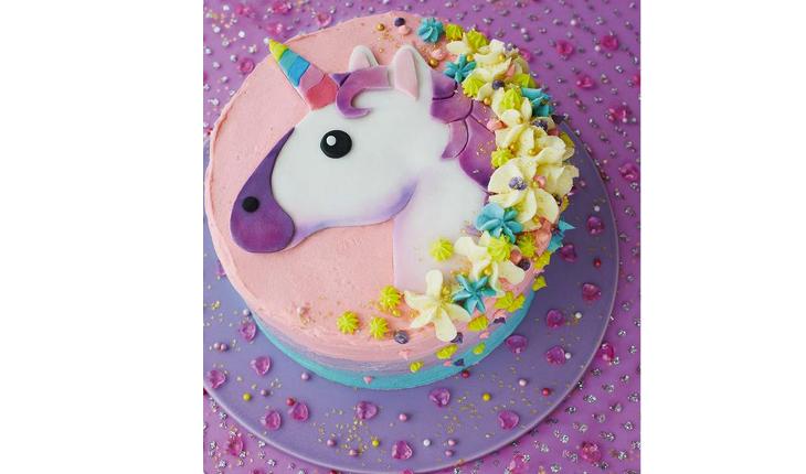 Na foto há um bolo redondo de unicórnio. No topo do bolo há um desenho de um unicórnio branco com detalhes roxos e chifre colorido. Ao redor do desenho há detalhes.