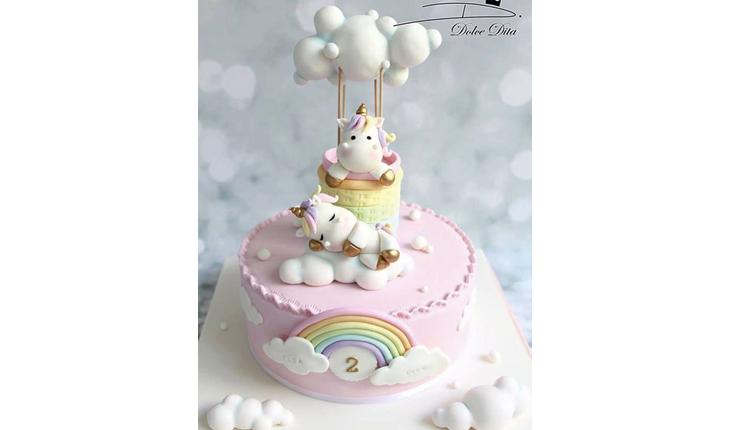 Na foto há um bolo redondo nos tons rosa-claro e com um arco-íris desenhado na frente. Em cima do bolo há dois unicórnios pequenos dormindo.