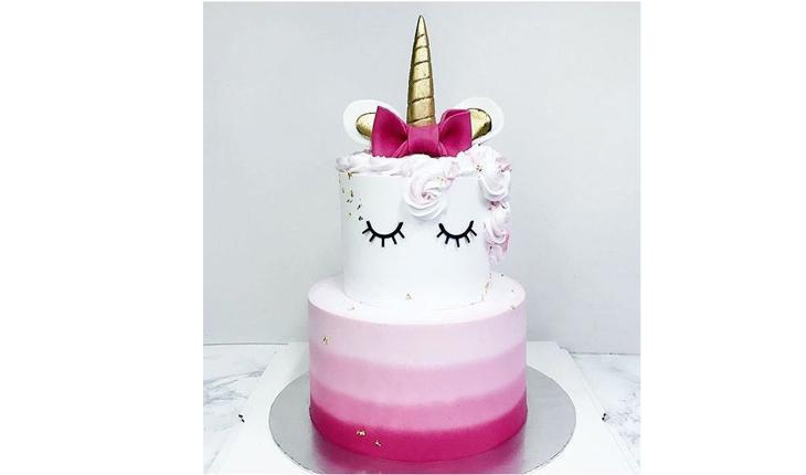 Na foto há um bolo de dois andares de unicórnio nas cores rosa e branca. O bolo de base tem listras rosas em degradê, sendo a mais escura no bolo de base. O bolo do topo é branco e tem os olhos do unicórnio desenhados. A crina é rosa-claro e o chifre é dourado. Ao redor do chifre há um laço pink.
