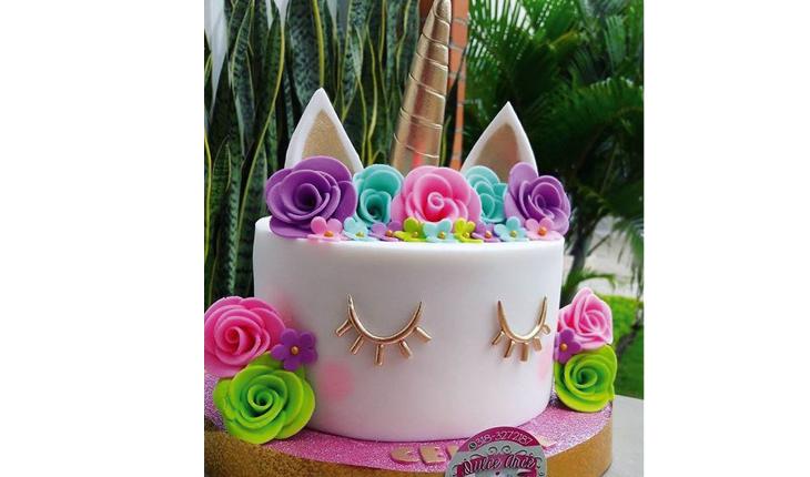Na foto há um bolo de unicórnio branco com detalhes em dourado, assim como o chifre. Ao redor e a crina é feita com flores nas cores verde, rosa e roxo.