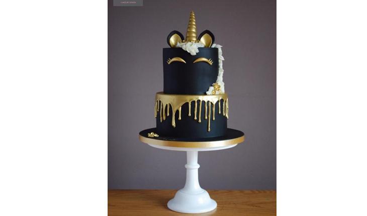 Na foto há um bolo de dois andares preto com detalhes em dourado e branco. A crina é branca e está no topo do bolo e cai ao redor. No topo há o chifre dourado. O rosto é feito apenas do os olhos fechados do unicórnio.