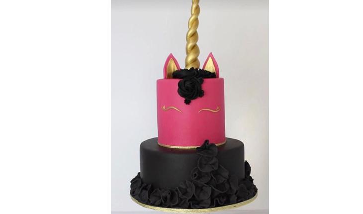 Na foto há um bolo de unicórnio de dois andares. O bolo de base é preto e o bolo do topo é rosa pink. A crina é preta e os detalhes são em dourado e o chifre também é dourado. O rosto é feito apenas do os olhos fechados do unicórnio.