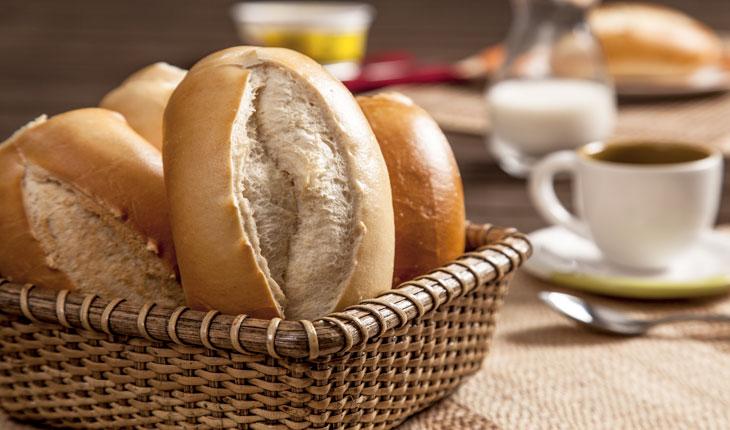 Na foto há um cesto com pães franceses em cima de uma mesa de café da manhã. Ao fundo há uma xícara.