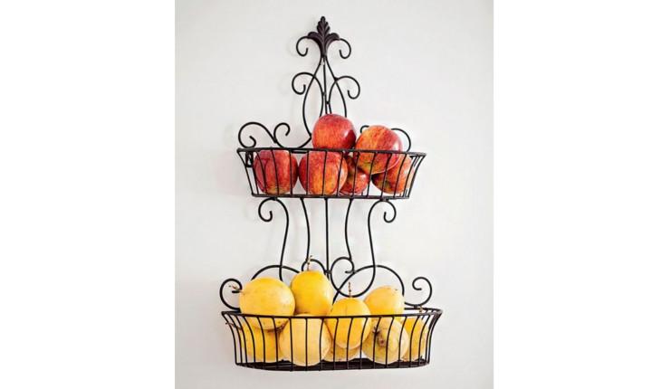 A foto mostra uma fruteira de metal decorado com duas partes. Na parte mais alta há uma fruta vermelha. Na parte mais baixa há uma fruta amarela. O suporte é pendurado na parede.
