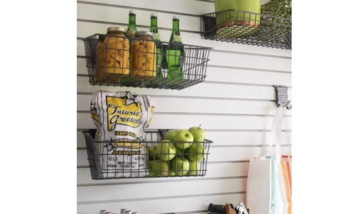 Na foto há diversos cestos de metal pendurados em uma parede branca com madeira. Em cada um dos cestos há alguns tipos de alimentos, como frutas e até garrafas de cerveja.