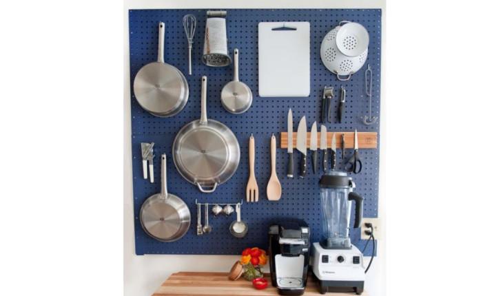 A foto mostra uma painel de metal pintado de zul em que os utensílios de cozinha, como panelas e colheres, ficam pendurados.