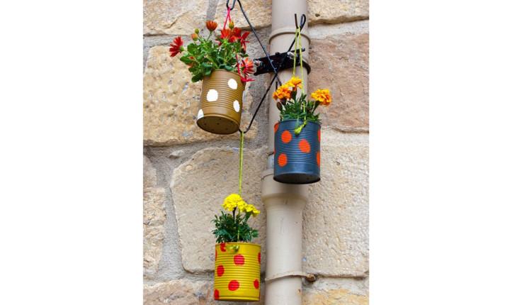 Na foto há latas de aço pintadas de cores vivas e decoradas com bolinhas de cores contrastantes também pintadas. As latas estão penduradas em um suporte de jardim e têm plantas dentro.