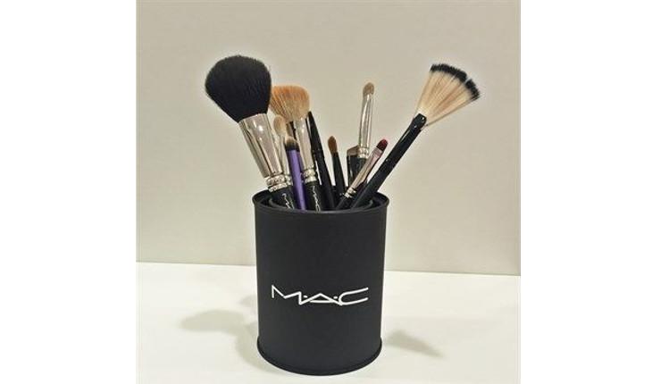 Na foto há uma lata pintada de preto e com a logo da marca MAC pintada em branco bem na frente. A lata está sendo utilizada para guardar pincéis de maquiagem.