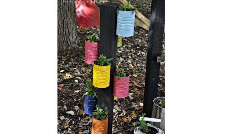 Na foto há um suporte de jardim em pé com latas pintadas de cores vivas presas ao redor. Cada uma delas há uma plantinha dentro.