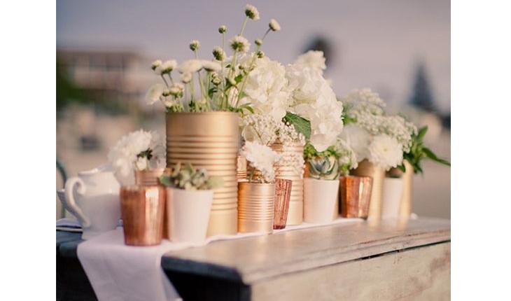 Na foto há várias latas de aço pintadas na cor cobre e com flores brancas dentro. O enfeite fica como um centro de mesa.