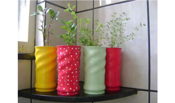 Na foto há um cantoneira de coziha com diversos vasinhos em cima. Os vasinhos são feitos com latas de achocolatado pintadas de cores vivas. Dentro de cada uma delas há um tipo de tempero plantado.