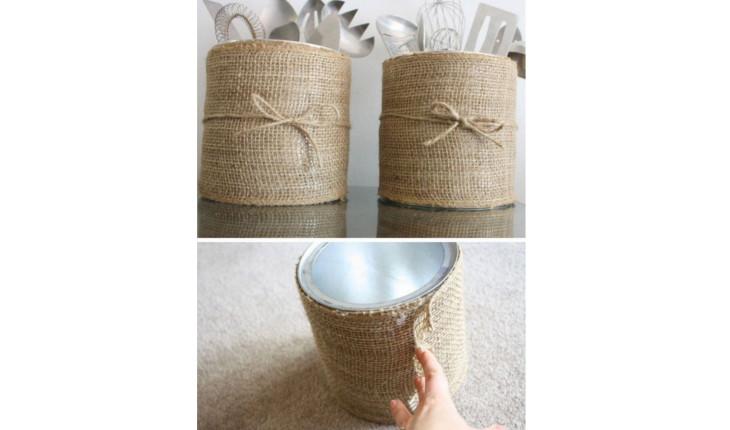 Na foto há uma lata de aço revestida com tecido bege e com um lacinho. A lata é utilizada para colocar talheres e decorar mesas.