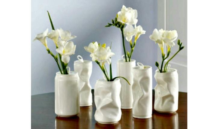Na foto há vasinhos feitos com lata de alumínio. As latas estão pintadas de branco e algumas são amassadas. Há flores brancas dentro de cada uma delas.