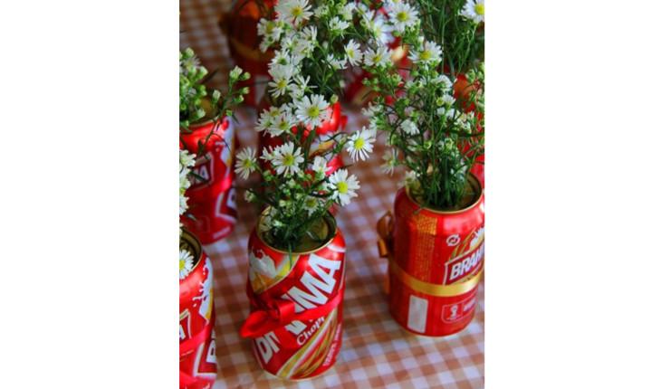 Na foto há diversas latinhas de cerveja na cor vermelha em que dentro de cada uma delas há plantinhas com flores brancas pequenas. Ao redor das latinhas há fitinhas vermelhas com laços. Esta é uma ideia de decoração de mesa para um chá-bar, por exemplo.