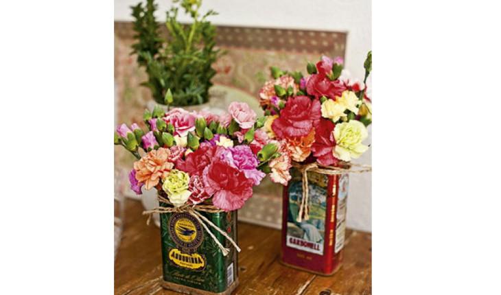 Na foto há 3 arranjos de flores feitos em latas de azeite. As latas estão em pé e servem como vasinhos. As latas são coloridas e as flores também.