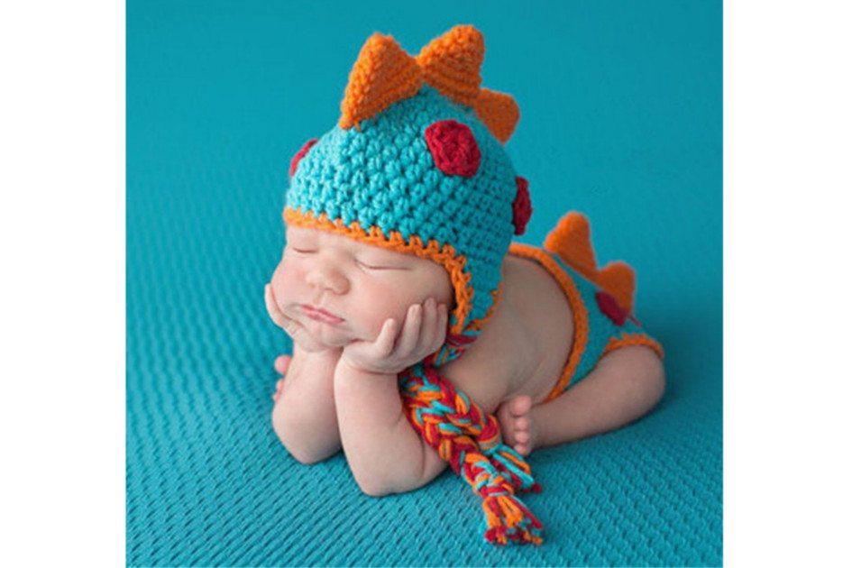 Na foto há uma fantasia de crochê para bebê de dinossauro na cor azul e laranja. Há um bebê recém-nascido vestindo a fantasia.