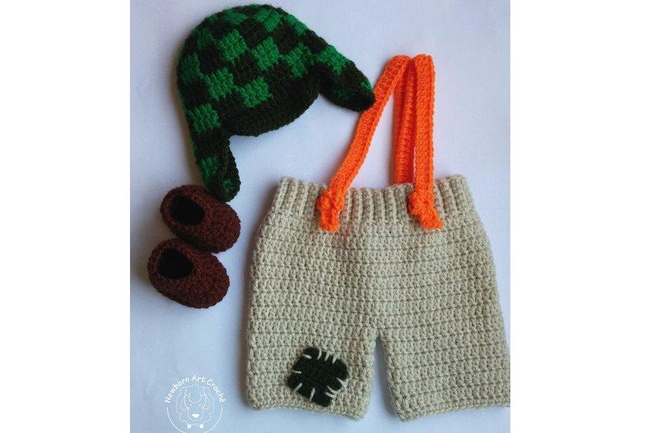 Na foto há uma fantasia do Chaves feita de crochê para bebês. O shorts é bege, há um suspensório laranja e um chapéu quadriculado, como do personagem, em verde e preto. Há também um sapatinho marrom.
