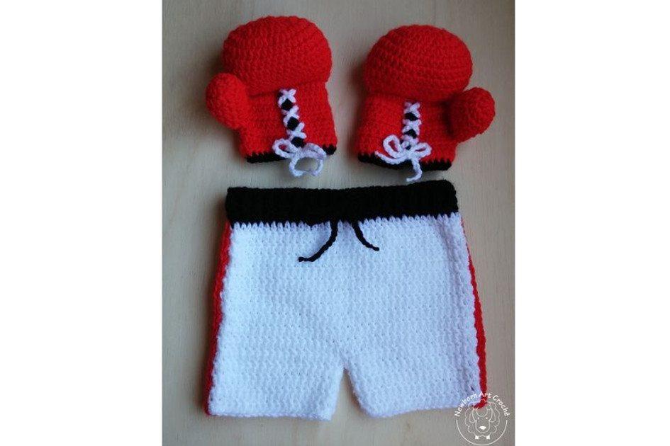 Na foto há uma fantasia de lutador de boxe feita de crochê. O shorts é branco e vermelho e a luva é é vermelha.