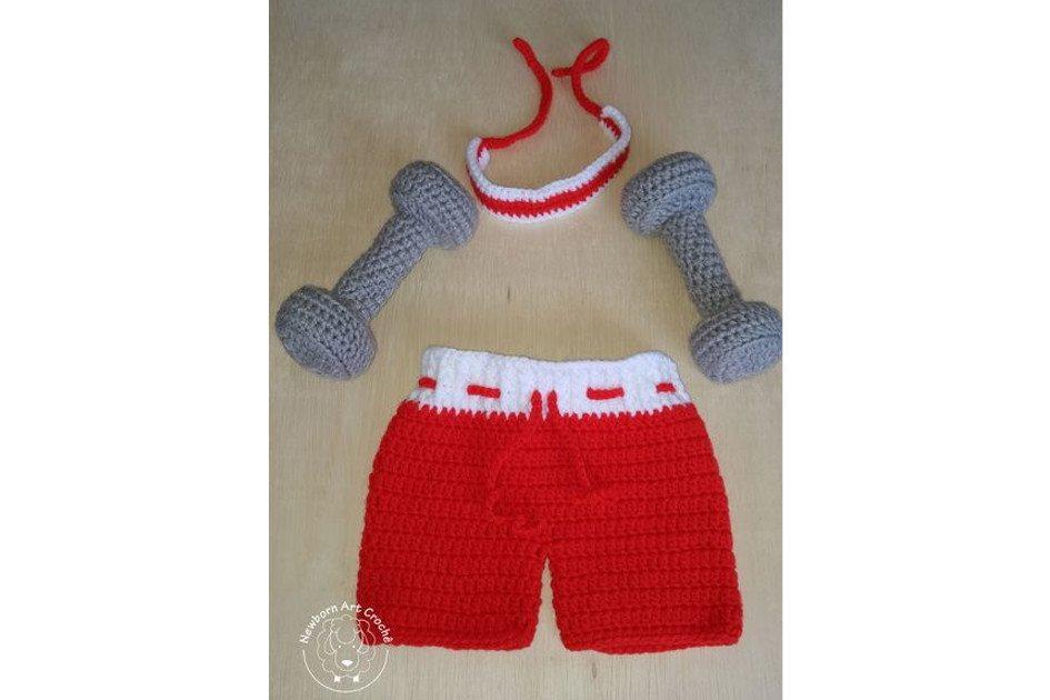 Na foto há uma fantasia de esportista feita de crochê com o shorts vermelho e os pesos em cinza.