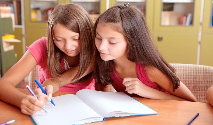 Na foto há duas meninas fazendo a tarefa de casa juntas, segurando uma caneta e olhando para um caderno.