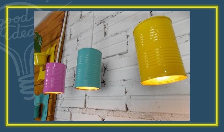 Na foto há 3 luminárias feitas de latas de aço pintadas de amarelo, rosa e azul-claro. Elas estão próximas a uma parede e as lâmpadas estão dentro delas.
