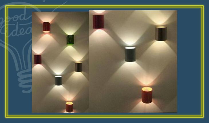Na foto há luminárias de parede feitas com latas de aço pintadas que são posicionadas de forma decorativa em uma parede. A luz sai pelas duas extremidades das latas.