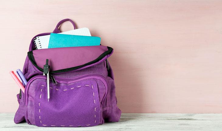 Na foto há uma mochila roxa com cadernos e outros materiais de escola.